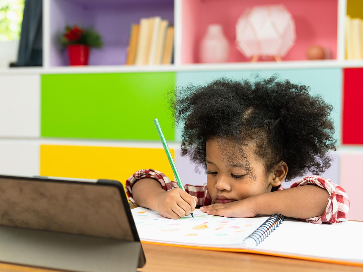 Girl sitting at a desk writing in a notebook and using a tablet