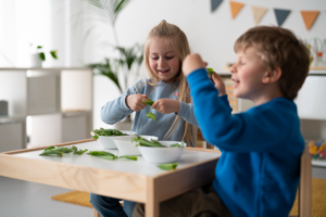 Two children sit at a classroom table, observing green leaves placed in bowls.