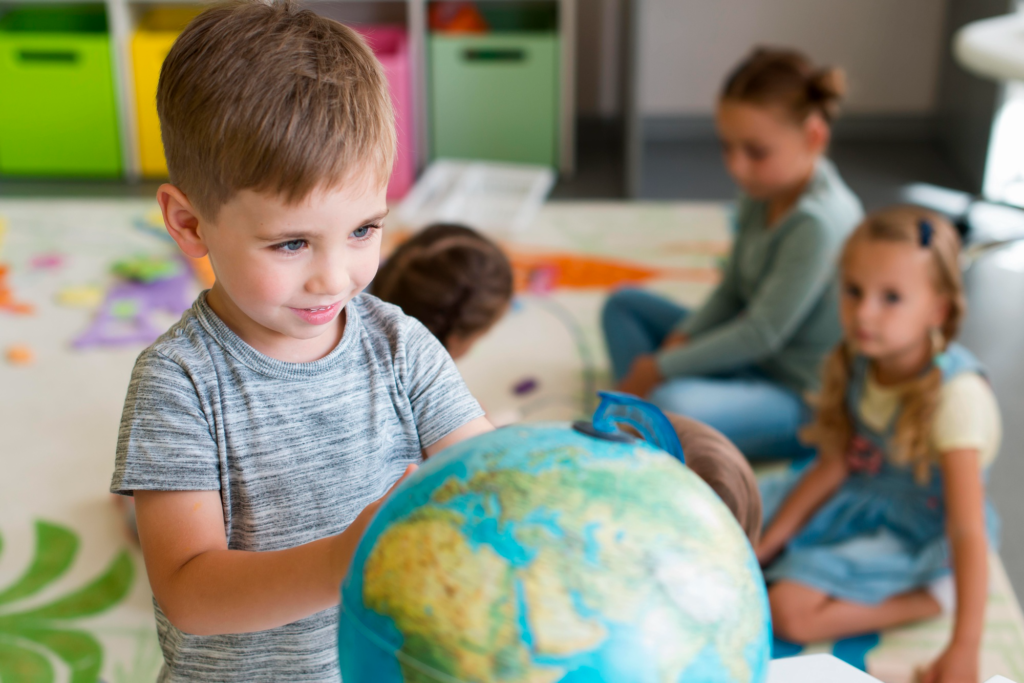 A young boy rotates a blue and green earth globe while sitting on a classroom rug.