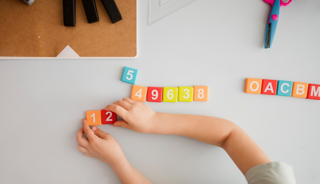 Overhead view of a child at a wooden desk practicing literacy and numeracy with physical letters and numbers.