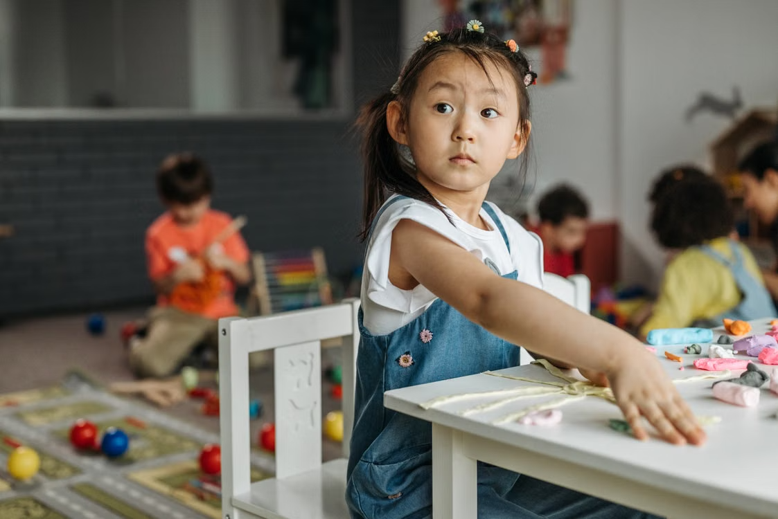 Young girl working with modeling clay while other children participate in activities in the background.